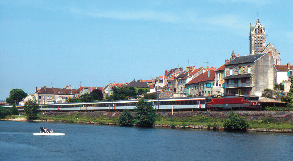 À Dormans, depuis la Marne, les occupants d’un pédalo admirent le passage de l’express Corail 1004 Strasbourg - Paris‑Est, au crochet de la BB 15009, filant en direction de la capitale (30 mai 1982 ; L. Chanteloup).
